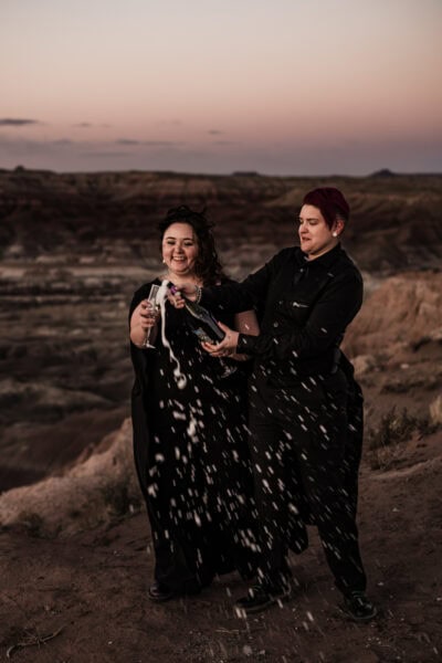 A joyous celebration in the Painted Desert, as the couple raises their glasses of champagne with close friends, the bride in a black wedding dress with a cape.