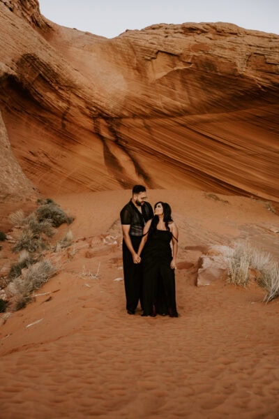 Surrounded by towering red rock formations, the couple shares a quiet moment of connection, their black attire contrasting beautifully with the desert landscape.