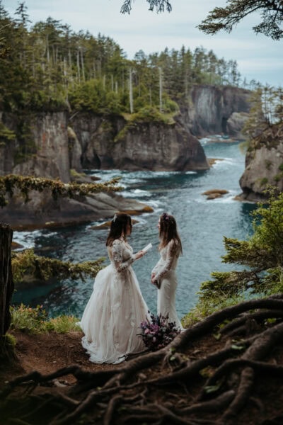 lgbtq elopement couple share private vows on an oceanside cliff overlooking the pacific ocean. towering seastacks and cliffs frame the blue water. they are surrounded by a lush forest that frames the edges of the cliffs.