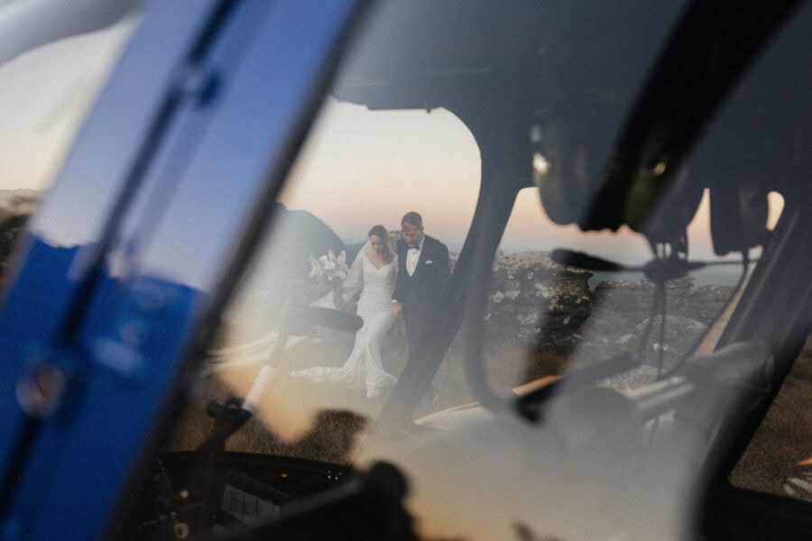 Bride and groom seen walking side by side through reflection of helicopter window during helicopter elopement in South Africa.